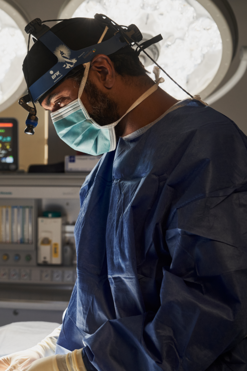 Dr. Paul Gill, wearing a blue surgical cap and mask, focuses intently during a surgical procedure in a well-equipped operating room.