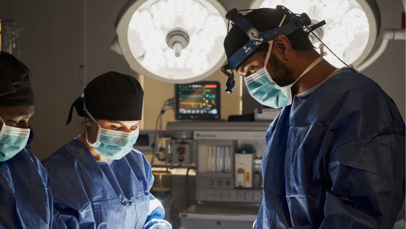 Dr. Paul Gill, wearing a blue surgical cap and mask, focuses intently during a surgical procedure in a well-equipped operating room.